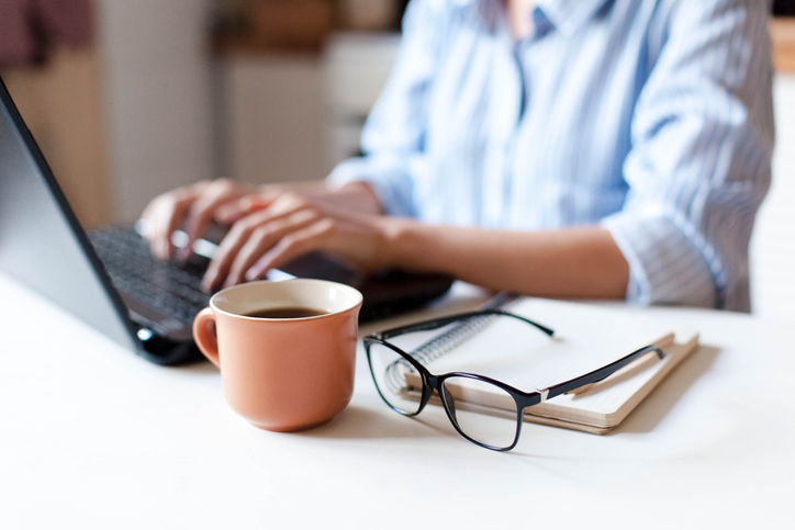 A person typing at a laptop with a coffee mug, glasses, and a note pad on their desk